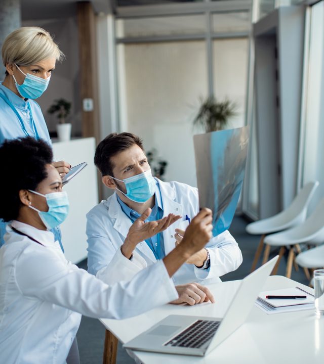 Group of doctors cooperating while examining X-ray of a patient Team of medical experts with protective face masks analyzing X-ray image at the clinic.