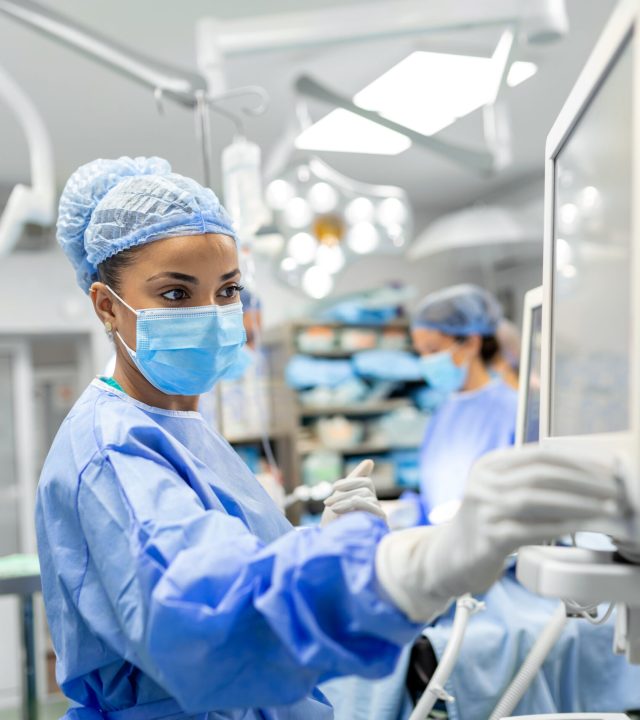 Anesthetist Working In Operating Theatre Wearing Protecive Gear checking monitors while sedating patient before surgical procedure in hospital Anesthetist Working In Operating Theatre Wearing Protecive Gear checking monitors while sedating patient before surgical procedure in hospital